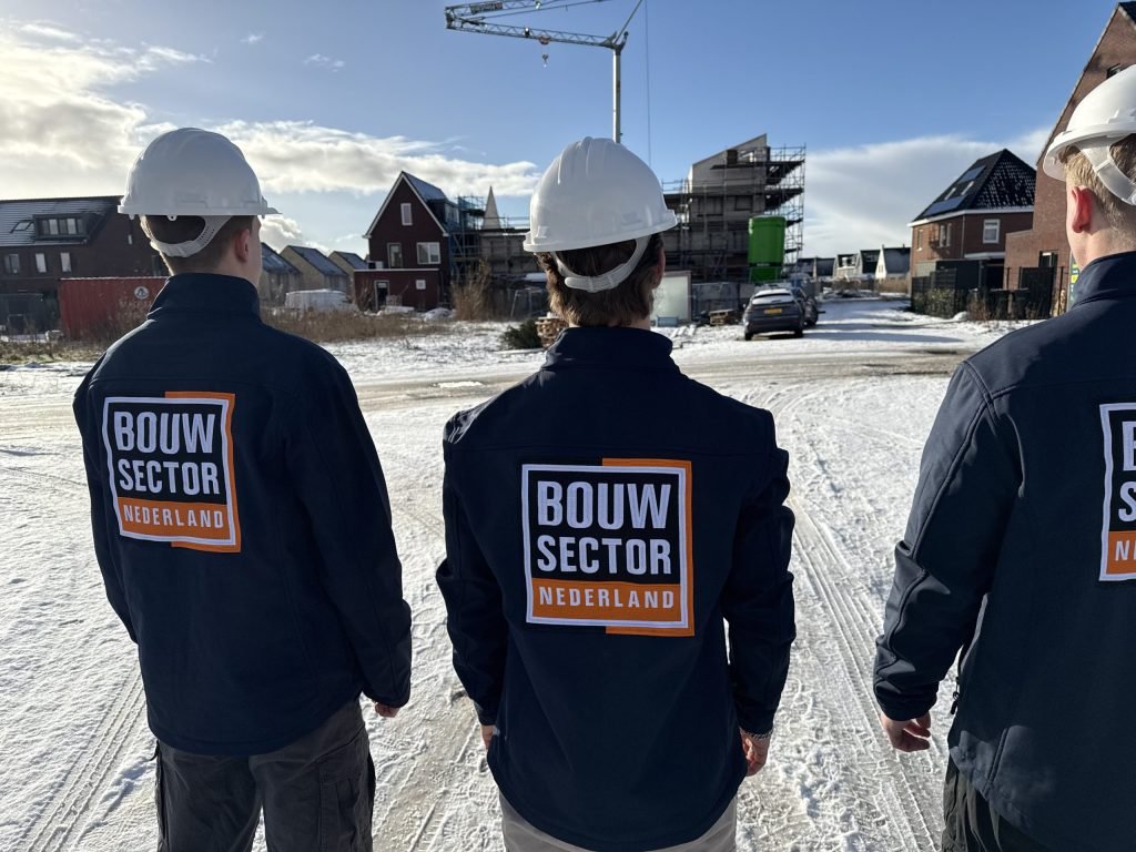 Drie vakmensen van Bouwsector Nederland met witte helm kijken over een besneeuwde straat met nieuwbouwhuizen en een bouwkraan