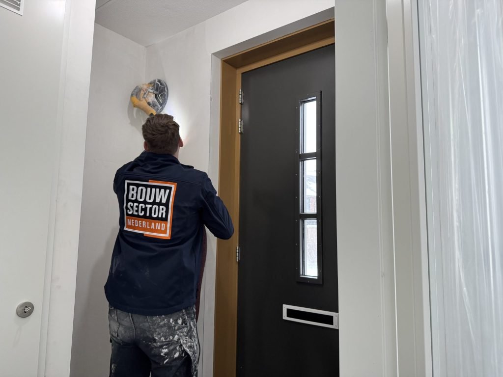 Vakman van Bouwsector Nederland in donkere jas schuurt boven een voordeur met een wandschuurmachine