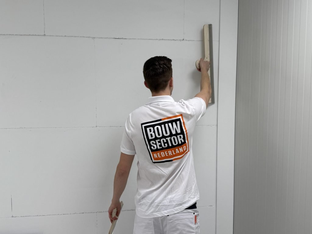 Stukadoor met het logo van Bouwsector Nederland op zijn rug zet een lange spaan verticaal tegen een wand